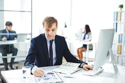 Portrait of young businessman busy working at desk in modern office: checking statistics report and using computer with colleagues in background