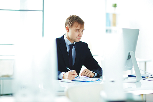 Portrait of successful young business analyst busy working alone in modern office, checking statistics report with graphs while sitting at computer desk