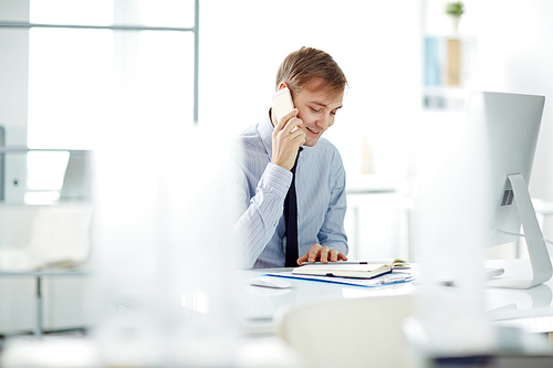 Portrait of successful young businessman wearing shirt and tie busy working alone in modern office, making calls using smartphone and smiling cheerfully  while sitting at computer desk