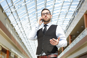 Low angle portrait of successful entrepreneur standing speaking by phone under glass ceiling in modern office building