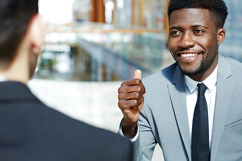 Portrait of young successful African-American businessman smiling and showing thumbs up while talking to partner, celebrating deal modern office building