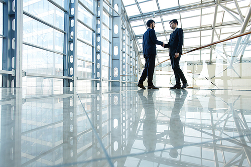 Wide angle portrait of two contemporary business men shaking hands in hall of futuristic glass building