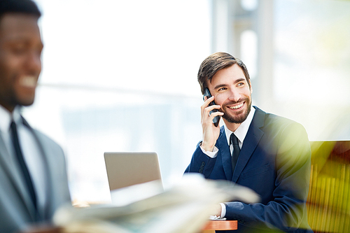 Portrait of smiling handsome businessman speaking on mobile phone while sitting at table in waiting lounge