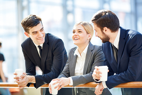 Portrait of three business people, one woman and two men, smiling cheerfully while chatting in glass hall of modern office building leaning on railing and holding coffee cups