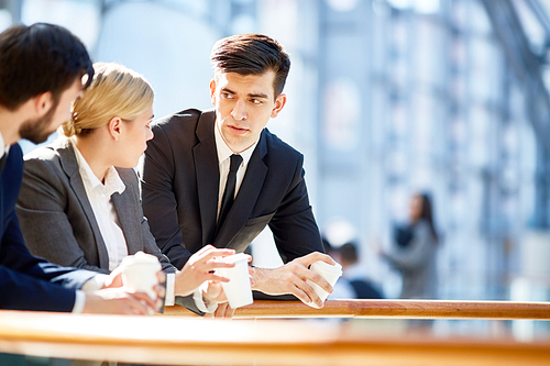 Portrait of three business people, one woman and two men, discussing work at break  in modern office building leaning on railing and holding coffee cups