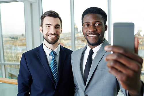 Portrait of handsome bearded businessman and his African partner taking smartphone selfie smiling to camera