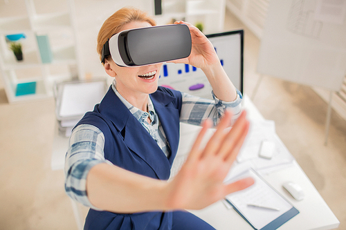 High angle view of cheerful middle-aged interior designer using VR headset while working on promising project, interior of spacious office on background