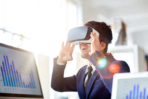Cheerful young businessman in stylish suit using VR headset while sitting at desk in modern open plan office, lens flare