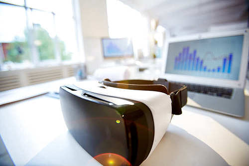 Close-up shot of VR headset lying on wooden desk, interior of spacious open plan office on background, lens flare
