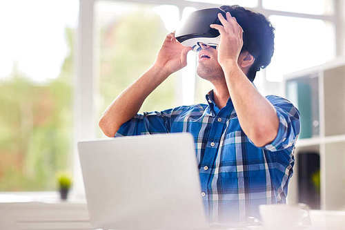 Young interior designer in checked shirt using VR headset in order to work with 3d visualization while sitting at desk in modern office
