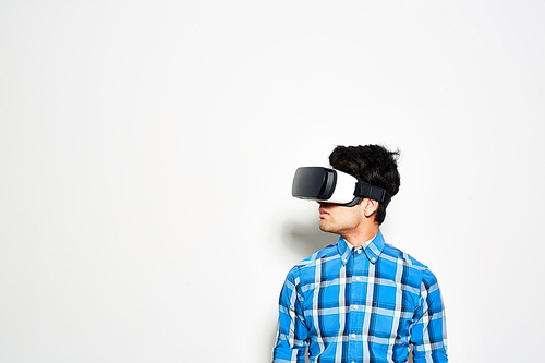Dark-haired young man looking away while testing virtual reality headset, white background
