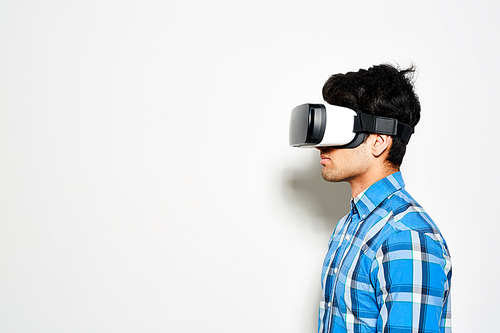 Profile view of confident young man using VR goggles while standing against white background, portrait shot