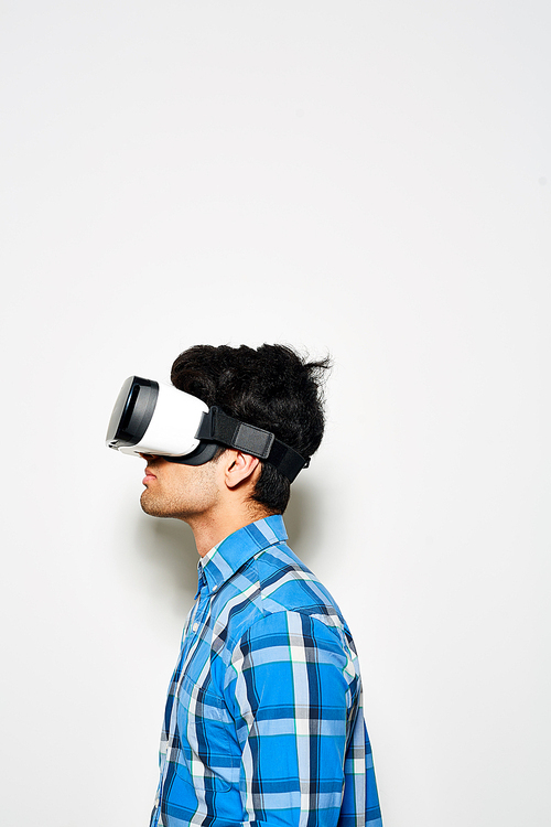 Concentrated young man in buttoned-up checked shirt using VR headset while standing against white background, profile view