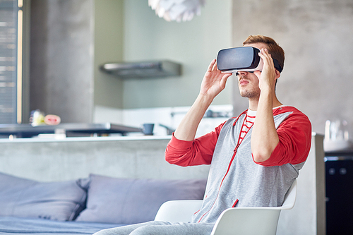 Portrait of young bearded man using VR headset while playing game, interior of modern studio apartment on background
