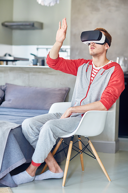Full length portrait of young man wearing sports suit sitting on cozy chair and using VR headset, interior of modern studio apartment on background