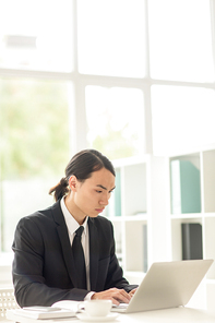 Confident Asian businessman wearing classical suit sitting in front of laptop and working on promising project, interior of modern office with panoramic windows on background