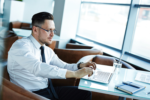 Waist-up portrait of confident middle-aged businessman working on presentation with help of modern laptop while sitting in office lobby, profile view