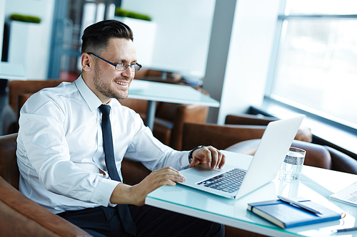 Waist-up portrait of smiling manager working on financial report with help of laptop while sitting in spacious office lobby with panoramic windows