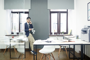 Cheerful young manager enjoying fragrant coffee and talking to his friend on smartphone while taking short break from work, interior of modern open plan office on background