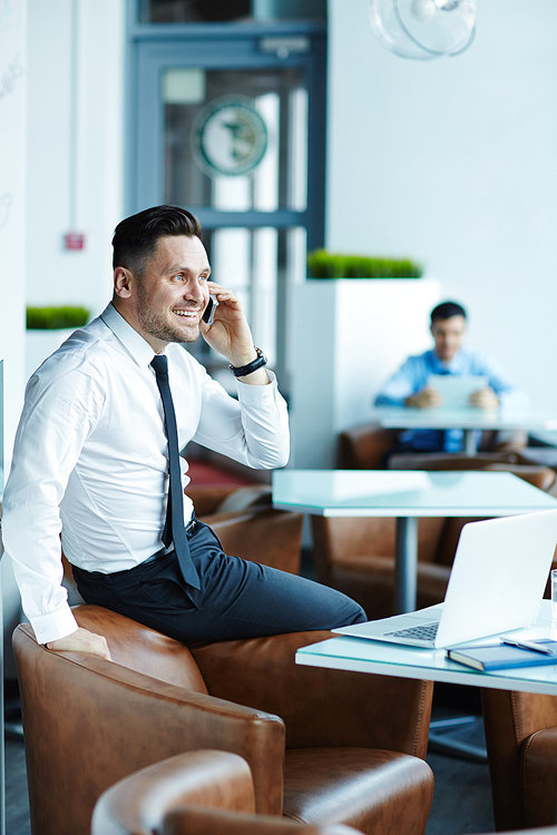 Joyful middle-aged businessman with stylish haircut talking to his colleague on smartphone while sitting in modern office lobby