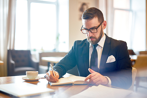 Busy director or ceo in suit sitting by table in cafe and writing in notebook