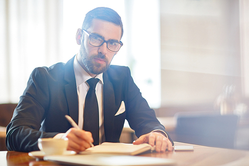 Young confident employer in suit making notes in notebook while planning working day