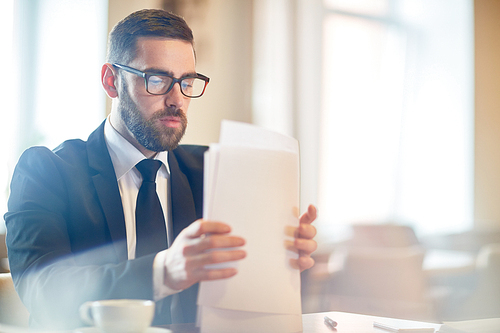 Confident bearded manager in classical suit sitting at office desk and doing paperwork, waist-up portrait