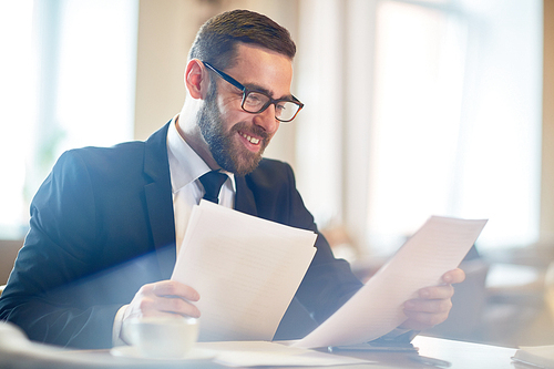 Cheerful young manager in eyeglasses analyzing results of accomplished work while sitting at office desk, blurred background