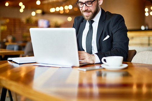 Smiling bearded manager in eyeglasses checking business emails on laptop while working at spacious restaurant, waist-up portrait