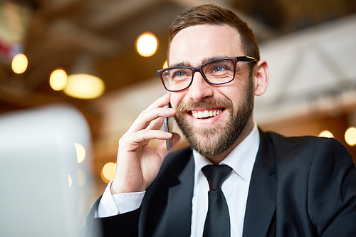 Joyful young businessman discussing joint project with colleague on smartphone while sitting at modern office, head and shoulders portrait