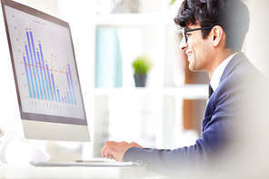 Profile view of smiling young manager in eyeglasses sitting at desk and preparing financial accounts, interior of modern open plan office on background