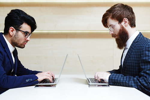 Profile view of two concentrated bearded businessmen in eyeglasses and suits sitting opposite one another and working on their laptops in office lobby, smartphones lying on desk