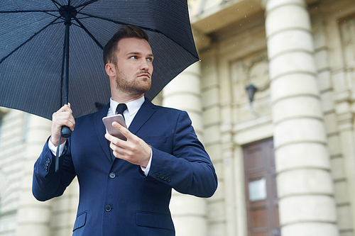 Business leader with smartphone waiting for somebody under umbrella on rainy day