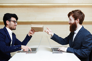 Birthday wishes: two bearded businessmen in eyeglasses working on laptops in office, one of them distracting another for a second in order to give him gift box without decorations