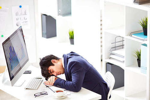 Overworked and tired young manager in stylish suit sleeping on desk in front of computer, interior of modern office on background