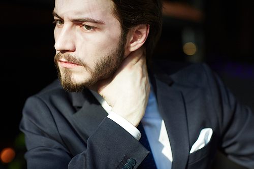 Sunlit head and shoulders portrait of handsome bearded man wearing elegant business suit, looking away frowning and rubbing his neck