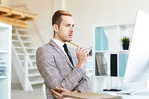 Profile view of pensive young financial manager with stylish haircut preparing annual accounts while sitting in front of computer at modern spacious office