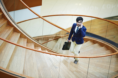 Portrait of modern middle ? eastern businessman wearing stylish formal suit going up spiral staircase in designer office building calling by smartphone