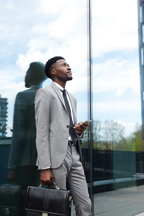 Young inspired African-american businessman with smartphone and briefcase leaning against wall of modern glassy building
