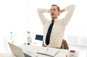 Handsome bearded businessman relaxing on office chair with hands behind head and feeling satisfaction of accomplished work, interior of modern open plan office on background