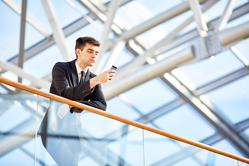 Young agent with smartphone messaging while leaning against railing