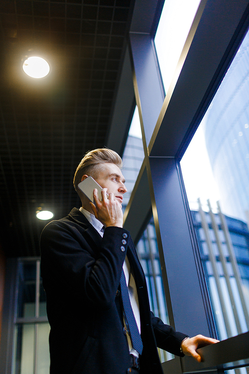 Low angle view of confident manager calling to his client on smartphone while standing by window in modern office lobby, profile portrait shot