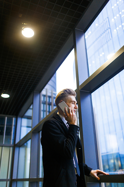 Profile portrait shot of young employee with stylish haircut talking to his business partner on phone while standing by panoramic window