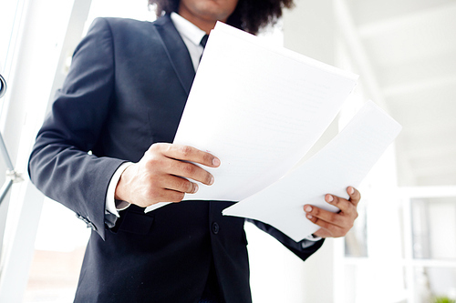 Low angle view of unrecognizable manager wearing suit studying documents while standing at open plan office, close-up shot