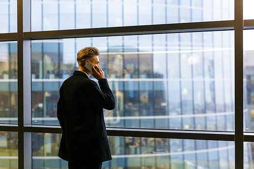 Back view of young businessman wearing suit standing at panoramic window while having project discussion with his colleague on smartphone