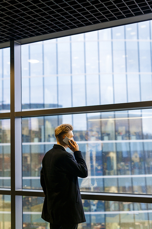 Unrecognizable businessman with stylish haircut looking out office window while discussing details of joint work with his colleague on phone, back view