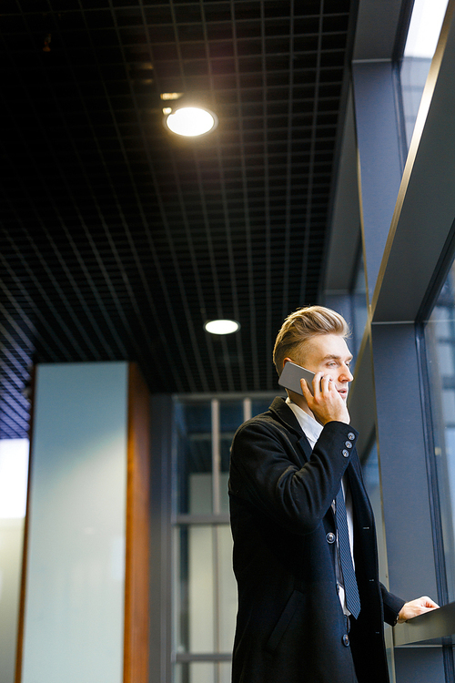 Thoughtful blond-haired white collar worker using smartphone while standing in office lobby and enjoying view from panoramic window, profile view