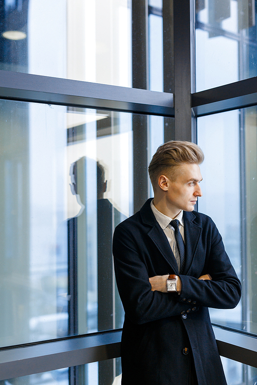 Thoughtful businessman wearing suit looking out window while standing with arms crossed in office corridor, waist-up portrait