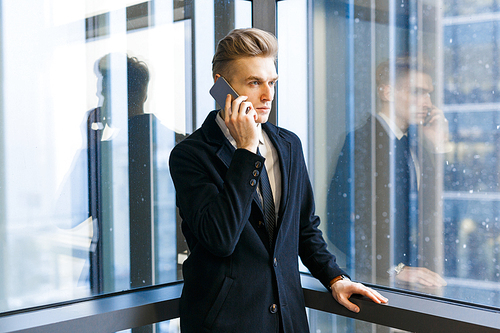 Portrait shot of serious white collar worker answering phone call while standing in office lobby with panoramic windows