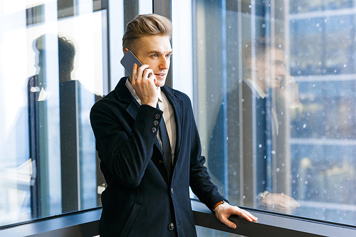 Young businessman with stylish haircut talking on phone while enjoying picturesque view from panoramic window of office lobby, waist-up portrait